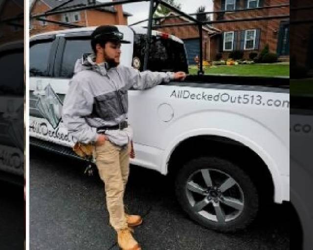 A man wearing work clothes and boots stands beside a white pickup truck with a business logo and the website AllDeckedOut513.com on the side. Houses and a lawn are visible in the background.