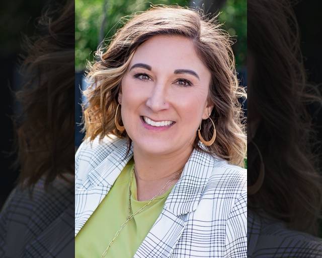 A woman with wavy, shoulder-length light brown hair smiles outdoors. She is wearing a white plaid blazer over a light green top and gold hoop earrings, with a blurred green background.