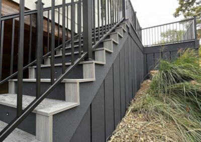 A modern outdoor staircase with black railings and gray steps leads up to a deck. Below, there is a rock and plant landscape beside a dark vertical-paneled wall. The sky above is overcast.