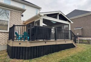 A modern backyard patio with a covered porch, black railing, and beige trim. Turquoise chairs sit on the deck, attached to a light brick house with large windows. Neighboring houses are visible in the background.