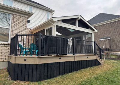 A modern backyard patio with a covered porch, black railing, and beige trim. Turquoise chairs sit on the deck, attached to a light brick house with large windows. Neighboring houses are visible in the background.