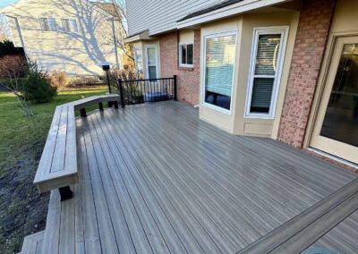 A modern, clean wooden backyard deck attached to a tan house with brick accents, featuring built-in bench seating and a small round table, surrounded by a grassy yard.