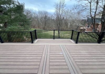 A spacious wooden deck with black railings overlooks a grassy backyard bordered by leafless trees. A house is partially visible on the right, and the sky is cloudy.