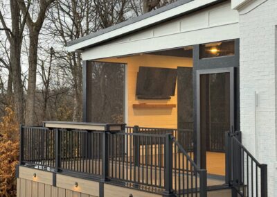 A modern screened porch with a white exterior, black railings, and wood-paneled steps, surrounded by leafless trees. The porch features a wall-mounted TV and built-in lighting along the steps.
