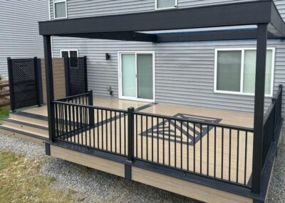 A modern backyard deck with black railings and geometric black designs on the wood floor, attached to a gray house with sliding glass doors and a small set of stairs on the left side.