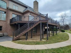 A two-story brick house with a large raised wooden deck featuring black railings and a long staircase leading to the backyard. The deck is supported by posts, and a curved sidewalk runs alongside the grassy yard below.