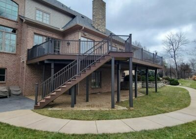 A two-story brick house with a large raised wooden deck featuring black railings and a long staircase leading to the backyard. The deck is supported by posts, and a curved sidewalk runs alongside the grassy yard below.