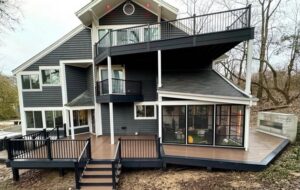 A modern gray house with multiple decks and balconies, black railings, and large windows. The house is surrounded by bare trees and has a tiered outdoor area with steps leading to the ground.
