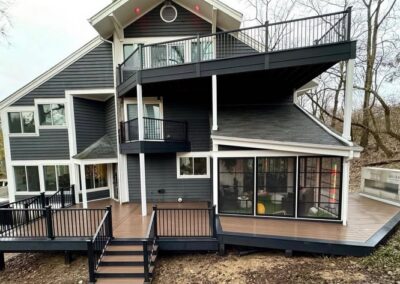 A modern gray house with multiple decks and balconies, black railings, and large windows. The house is surrounded by bare trees and has a tiered outdoor area with steps leading to the ground.
