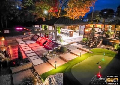 A modern backyard at night features illuminated landscaping, putting green, stone paths, a covered outdoor bar and seating area, and glowing lights; a National Deck Competition 2025 second-place badge is visible.