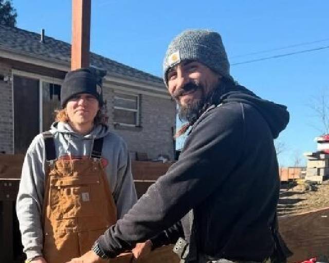 Two people stand outside on a sunny day; one wears brown overalls and a black beanie, the other wears a dark jacket and a gray knit hat, smiling at the camera. A house and construction materials are visible in the background.